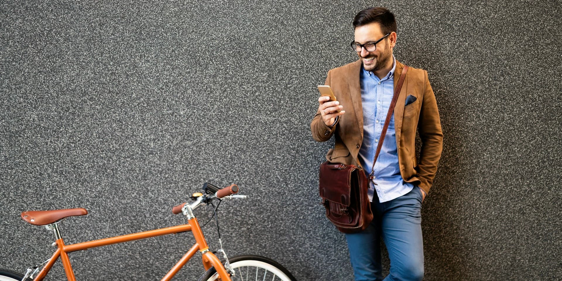 En un artículo sobre cuál es más barato para la caída del cabello en línea, un hombre vestido de negocios informal está de pie junto a una pared, sonriendo mientras mira su teléfono, con una bicicleta apoyada cerca.